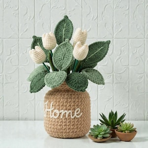 May include: A crocheted beige vase with the word "Home" in white script holds a bouquet of crocheted white tulips and green leaves. Small potted succulents sit nearby. The vase is approximately 8 inches tall.
