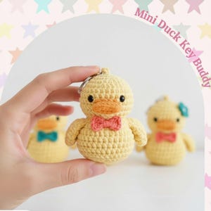 May include: A yellow crocheted duck keyring with a red bow tie and orange beak is held. The text "Mini Duck Key Buddy" is visible. Two other duck keyrings are in the background, one with a blue bow.