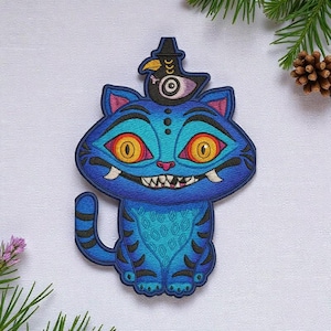 May include: Embroidered patch featuring a blue cat with large yellow eyes and sharp teeth. The cat wears a hat with a bird on top. The patch has intricate details and a whimsical design, perfect for adding to clothing or accessories.