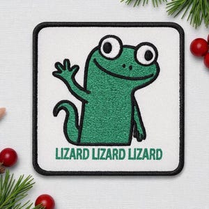 May include: Square patch with a green lizard waving, embroidered on a white background. The patch has a black border and the text "LIZARD LIZARD LIZARD" in green. The lizard has large eyes and a smiling expression.