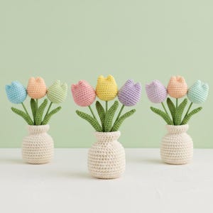 Può includere: Tre piccoli vasi, ognuno con un bouquet di tulipani all'uncinetto. I tulipani sono in vari colori pastello: blu, pesca, verde, rosa, giallo e viola. I vasi sono color crema, su uno sfondo verde chiaro.