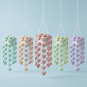 Puede incluir: Cinco macetas colgantes de ganchillo en colores pastel: verde menta, melocotón, rosa, verde claro y lavanda. Cada maceta es blanca con una guirnalda de corazones de ganchillo. Las macetas están suspendidas por cuerdas blancas sobre un fondo azul claro.