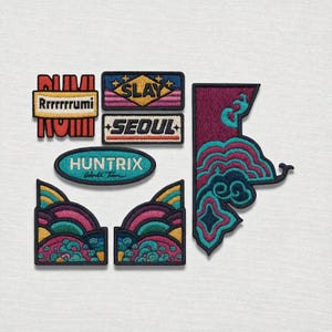 May include: A collection of embroidered patches in various shapes and designs. The patches feature text like "SLAY", "SEOUL", and "HUNTRIX". Colors include teal, pink, yellow, and purple. The patches are likely for clothing or accessories.