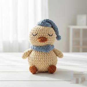 May include: A crocheted duck toy in a sleeping pose, wearing a blue hat and scarf. The duck has a beige body, a brown beak and feet, and closed eyes. The toy is handmade and sits on a white surface.