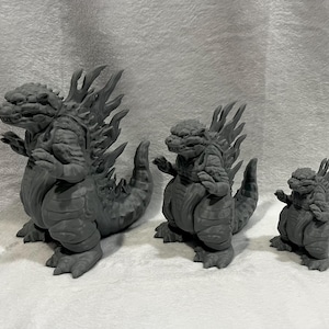 May include: Three gray Godzilla figurines of varying sizes are displayed. The largest figure is in the front, with two smaller versions behind it. Each figure has a detailed, textured design, including dorsal fins and a distinctive tail.