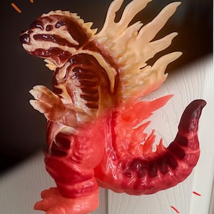 May include: A detailed sculpture of a monster with a red, orange, and brown color scheme. The creature has a spiky back, a long tail, and a detailed head. The sculpture is posed as if it is standing. The sculpture is on a white surface.