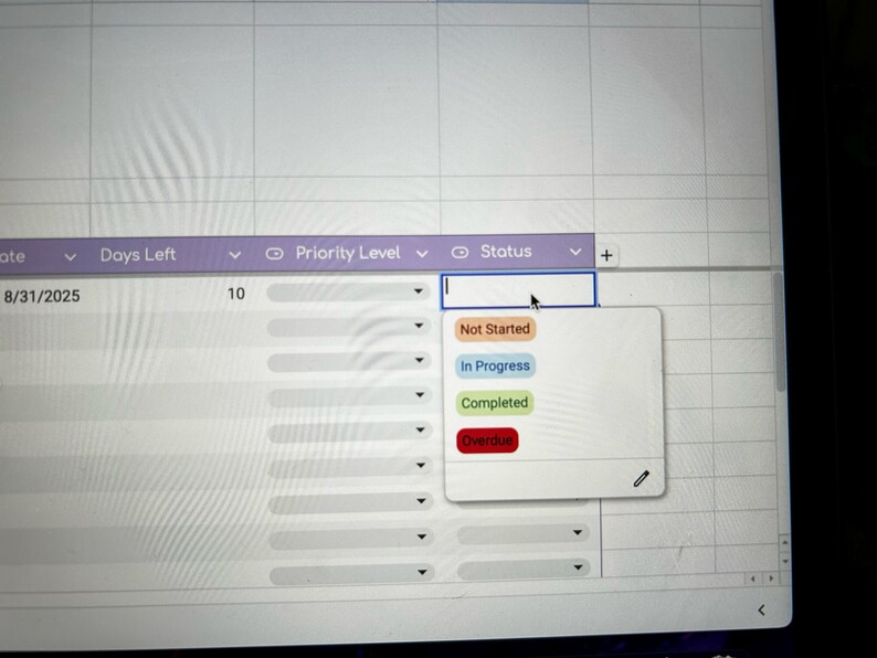 May include: A computer screen displaying a spreadsheet interface. Columns include 'Date', 'Days Left', 'Priority Level', and 'Status'. A dropdown menu offers status options: 'Not Started', 'In Progress', 'Completed', and 'Overdue'.