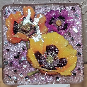 May include: A decorative night light with a square resin top, showcasing dried flowers in orange, yellow, and purple. The resin is filled with glitter and sits on a white plug-in base. The night light is designed for indoor use.