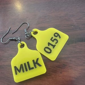 May include: A pair of yellow earrings shaped like livestock tags. One earring reads "MILK" in black text, and the other reads "0159". The earrings have silver-colored fish hook closures.