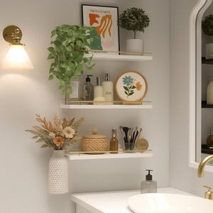 May include: A bathroom scene with three white shelves adorned with decorative items. A framed art print, plants, embroidered art, and toiletries are displayed. A white vase holds dried flowers, and a gold-toned sconce provides light.