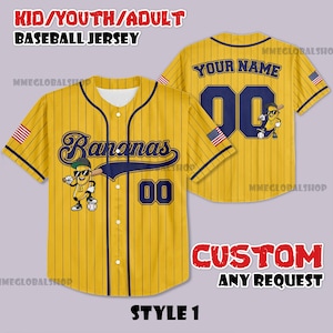 Puede incluir: Camiseta de béisbol amarilla con finas rayas azul marino. La parte delantera presenta la palabra "Bananas" en escritura azul marino, un personaje de plátano de dibujos animados y el número "00". La parte trasera tiene "YOUR NAME" y el número "00". Incluye detalles de la bandera americana.