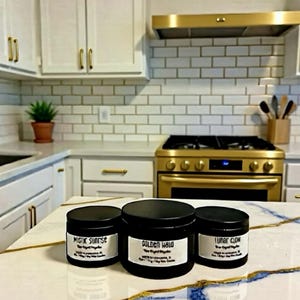 Può includere: Tre barattoli di candele neri con etichette bianche su un piano di lavoro in marmo. Le etichette recitano "Mystic Sunrise", "Golden Halo" e "Lunar Glow". La cucina presenta mobili bianchi, un fornello dorato e un paraschizzi in piastrelle bianche.