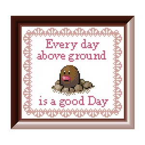 Może przedstawiać: Wzór haftu krzyżykowego przedstawiający brązową i różową postać Pokémona, Digletta, z tekstem "Every day above ground is a good day". Wzór jest oprawiony w brązową ramę z różowo-białym obramowaniem.