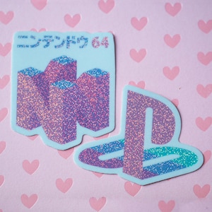 May include: Two glitter stickers on a pink background with heart shapes. One is light blue with Japanese text and purple shapes. The other is a PlayStation logo with a gradient of purple, blue, and green.