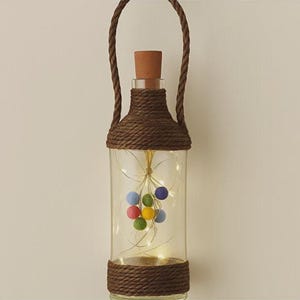 Puede incluir: Una linterna de botella de vidrio transparente envuelta en cuerda marrón oscuro con un asa de cuerda a juego y un tapón de corcho. Dentro, una cadena de luces ilumina un grupo de pequeñas bolas de fieltro de colores azul, verde, amarillo y rojo.