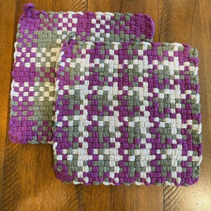 Puede incluir: Dos salvamanteles tejidos con un patrón geométrico. Los salvamanteles son principalmente morados, con detalles blancos y grises. Son cuadrados y parecen hechos a mano. Están sobre una superficie de madera.