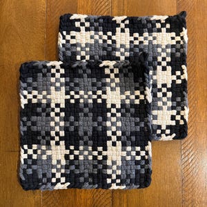 May include: Two woven potholders in a plaid pattern. The potholders are in shades of black, gray, and white. The potholders are square and rectangular. The potholders are made of a woven material.