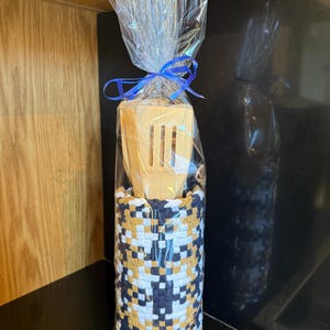 May include: A wooden spatula set in a woven holder, wrapped in clear cellophane and tied with a blue ribbon. The holder features a geometric pattern in navy, gold, and white. The spatula is made of light-coloured wood.