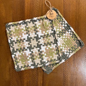 May include: Two square, woven potholders with a geometric pattern in shades of green, white, and beige. The potholders are handmade, as indicated by the attached tag. The potholders are on a wooden surface.