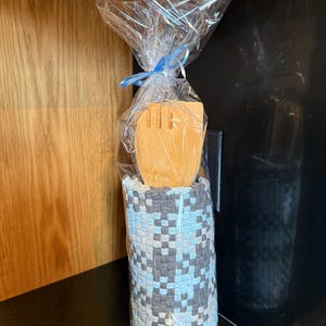 May include: A wooden spatula is packaged in clear cellophane, tied with a blue ribbon. It sits within a woven holder with a blue, grey, and white checkered pattern. The holder is cylindrical in shape, suitable for kitchen storage.