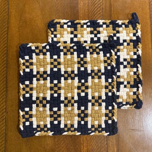 May include: Two square woven pot holders with a geometric pattern. The pattern features a grid of navy blue, cream, and gold squares. The pot holders have a navy blue border and a loop for hanging. The pot holders are made of a thick, durable material.