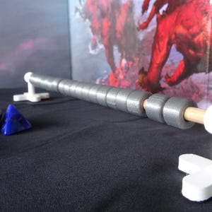 Puede incluir: Una herramienta de conteo gris y cilíndrica estilo ábaco con espiga de madera y soportes blancos. La herramienta de conteo está sobre una superficie negra con dados y un fondo de arte de fantasía. Una herramienta divertida para juegos de mesa.