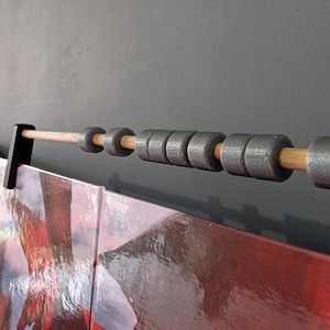 Może przedstawiać: Drewniany kołek z szarymi metalowymi pierścieniami, zawieszony na czarnym wsporniku. Kołek podtrzymuje dzieło sztuki w kolorach czerwonym i białym. Tło to jednolita szara ściana.