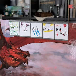 Może przedstawiać: Konfiguracja gry planszowej z ilustracją czerwonego smoka i kartami z imionami z napisami "Goblin", "The Dark Lord", "Bob", "Alice" i "Red Knight". Do planszy przymocowana jest mała czerwona flaga.