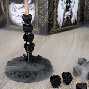 Puede incluir: Un porta incienso negro con temática de calaveras con una base decorativa y una pila de calaveras. El soporte está rodeado de humo y pequeños candelabros con forma de calavera. Una botella de líquido verde está en el fondo.