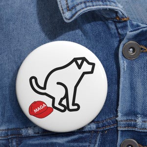 May include: White button with a black outline of a dog pooping, and a red baseball cap with the word "MAGA". The button is set against a blue denim background.