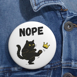 Puede incluir: Un botón blanco con la palabra "NOPE" en letras negras. Debajo, un gato negro pateando una corona dorada. El gato tiene ojos amarillos y una expresión de enfado.