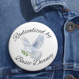 May include: A white button with the text "Radicalized by Basic Decency" in black script. The button features a watercolor illustration of a dove carrying an olive branch. The button is set against a blue denim background.