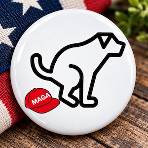 May include: A circular white button featuring a black line art illustration of a dog in a crouching position. Positioned below the dog is a red baseball cap with the white text MAGA printed on the front of the cap in a bold, sans-serif font.