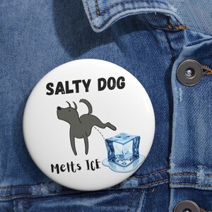 May include: A white button featuring a cartoon dog urinating on a melting ice cube. The text "SALTY DOG" is above the dog, and "Melts Ice" is below. The dog is gray, and the ice cube is blue.