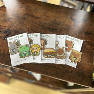 Puede incluir: Seis etiquetas de pan de madera con diseños de animales de dibujos animados y alimentos, incluyendo un conejo, una rana, un girasol, un sándwich, una mariquita, un oso y un búho. Cada etiqueta está empaquetada en una funda de plástico transparente. El texto "Bread Tag: Pack of 2" es visible.