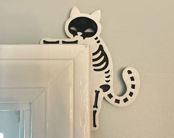 May include: A wooden cat skeleton decoration with a black face and rib cage, positioned on a white door frame. The cat's tail curves down, and the design is perfect for Halloween or spooky decor.