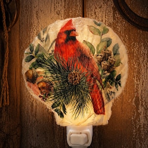 May include: A decorative night light featuring a vibrant red cardinal bird design. The bird is surrounded by green foliage and pine needles, set against a cream-colored, shell-shaped background. The night light has a white base with an on/off switch.