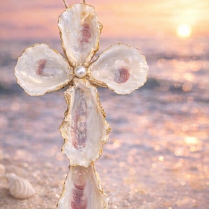 May include: A decorative cross made from oyster shells, with gold-painted edges and a pearl at the center. The cross is suspended by a natural twine loop. The background features a beach scene with a sunset.