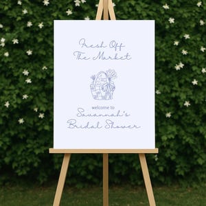 May include: A white sign on a wooden easel with the text "Fresh Off The Market" and "welcome to Savannah's Bridal Shower" in blue script. A blue illustration of a basket with vegetables is in the center.