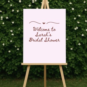 May include: A light pink sign on a wooden easel with the text "Welcome to Sarah's Bridal Shower" in a decorative script. The sign is decorated with a small heart and a curved line. The background is a green hedge.