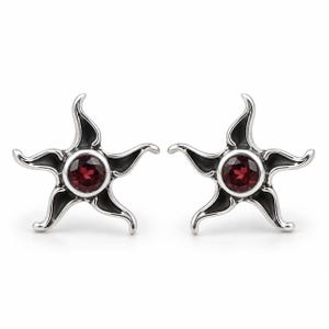 May include: A pair of silver-toned stud earrings shaped like five-pointed stars with curved, flowing arms. Each earring features a round, deep red gemstone set securely in the center, surrounded by dark, recessed detailing that highlights the metal form.
