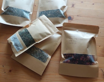 May include: Several beige paper pouches filled with dried herbs and flowers are arranged on a wooden surface. The pouches have handwritten labels. One pouch contains red rose petals. The other pouches contain green herbs.