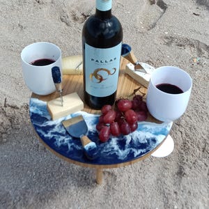 May include: A beach picnic setup featuring a bottle of wine, two white cups filled with red wine, cheese, grapes, and a serving board with a blue and white wave design. The wine bottle has a blue label with gold lettering.