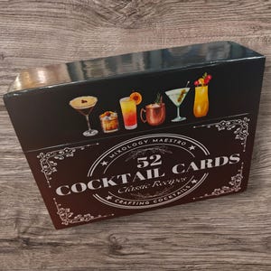 May include: A black box featuring illustrations of various cocktails. The box reads "Mixology Maestro 52 Cocktail Cards Classic Recipes Crafting Cocktails" in white text with decorative flourishes. The box is on a wooden surface.