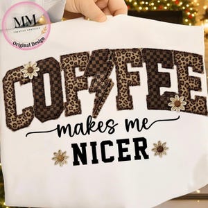 May include: White t-shirt with the text "COFFEE makes me NICER" in a bold, decorative font. The word "COFFEE" features a leopard print and checkered pattern, with a lightning bolt and flower accents. The shirt is a casual wear item.
