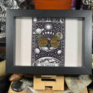 May include: A framed print with a butterfly and a celestial theme. The artwork shows planets, moons, and stars, with the words "THE UNIVERSE" at the bottom. The frame is black and the background is off-white.