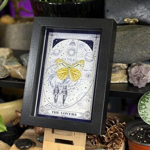 K&ouml;nnte beinhalten: Gerahmter Tarotkarten-Druck mit einem gelben Schmetterling und zwei H&auml;nden haltenden Figuren, betitelt "THE LOVERS". Das Kunstwerk befindet sich vor einem Himmels Hintergrund mit Planeten und Sternen, in einem schwarzen Rahmen.