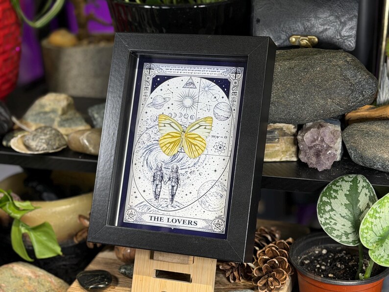 K&ouml;nnte beinhalten: Gerahmter Tarotkarten-Druck mit einem gelben Schmetterling und zwei H&auml;nden haltenden Figuren, betitelt "THE LOVERS". Das Kunstwerk befindet sich vor einem Himmels Hintergrund mit Planeten und Sternen, in einem schwarzen Rahmen.