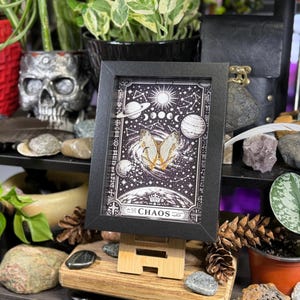 May include: A framed black and white print with celestial and butterfly imagery. The print features planets, moons, and stars, with the word "CHAOS" at the bottom. The frame is displayed on a wooden easel, surrounded by plants and decorative items.