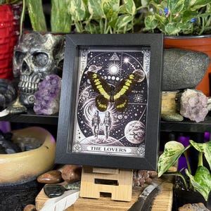 Puede incluir: Una impresi&oacute;n enmarcada en blanco y negro de la carta del tarot "Los Amantes", con una mariposa con alas amarillas y negras. La impresi&oacute;n se muestra en un caballete de madera, rodeada de plantas y objetos decorativos.